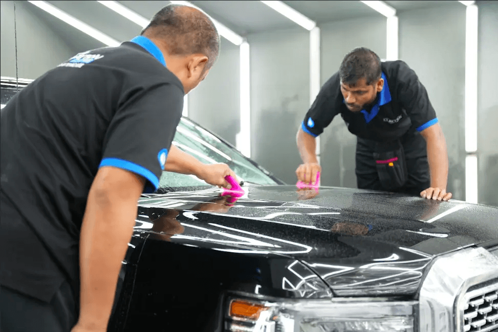 Two Clayton Care technicians applying paint protection film (PPF) to the hood of a black vehicle using pink squeegee tools in a well-lit detailing bay.